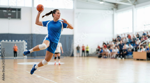 Dynamic female handball player jumping to throw the ball during a match in an indoor arena. Athletic woman in blue uniform performing a jump shot. Competitive sports and action concept
