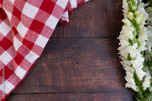 Red and white checkered cloth rests on dark wood. White flowers line the edge, delicate and fresh. . Perfect for rustic, garden, or picnic-themed projects
