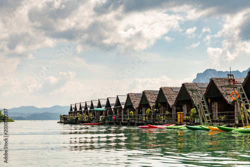 Tableau sur toile Serene row of wooden overwater bungalows with thatched roofs, solar panels, and colorful kayaks floating on calm turquoise water, against a backdrop of lush green mountains and dramatic cloudy sky