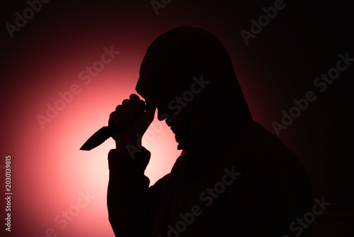 Silhouette of a hooded man holding a tactical knife against a dramatic red background, concept of crime, danger and thriller