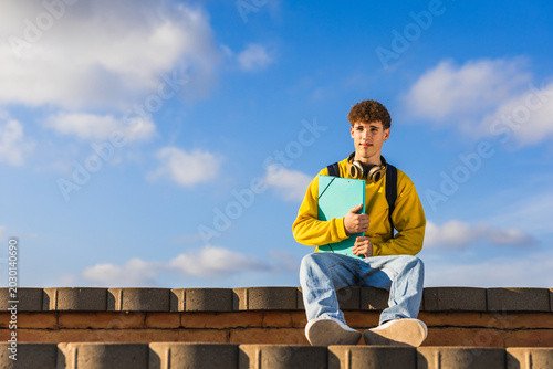 Young college student holding a binder sitting on a wall outdoors