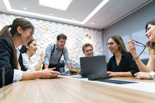A diverse group of young professionals gather around a wooden conference table, engaged in a discussion led by a standing presenter.