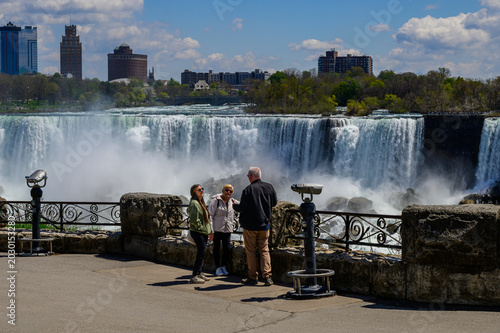 Fotografía Two tourists with guide viewing Niagara Falls from Canadian side observation area, spring, with American skyline visible in background