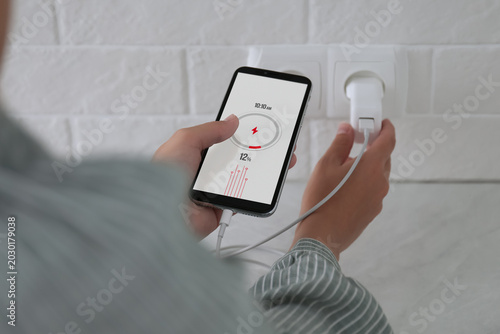 фотографія Woman with mobile phone plugging charger in socket on white brick wall, closeup