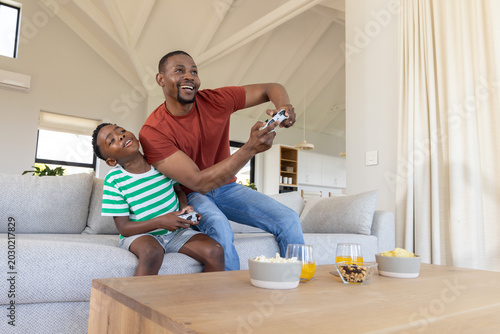 Картината върху платно African American dad and son on sofa holding controllers and snacking at coffee