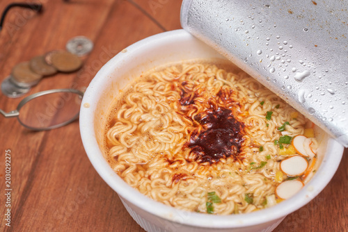 Close-up shots of Korean instant cup noodles on a wooden table. Wavy noodles in a spicy red broth are lifted by metal chopsticks. Blurred coins and glasses in the background suggest a quick meal.