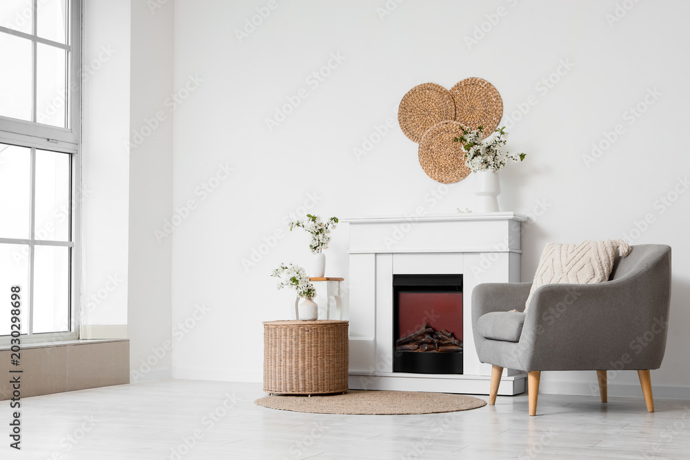 Fototapeta premium Interior of light living room with blooming branches in vases, fireplace and armchair