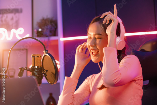 Bild auf Leinwand Host channel of young beautiful Asian broadcaster listening the music on social media with listeners, wearing pastel headphones, using microphone for singing at neon light indoor studio