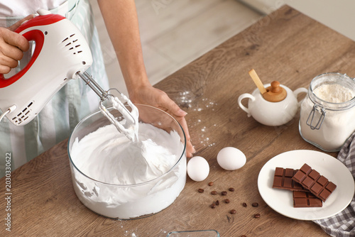 Obraz na plátne Woman whipping egg albumen with mixer at table in kitchen, closeup