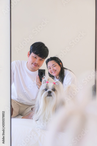 Young Asian couple and their dog taking mirror selfie together in cozy bedroom.