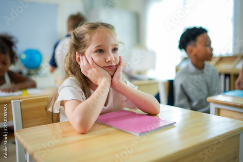 Thoughtful elementary schoolgirl resting face on hands at classroom desk during lesson while classmates continue studying in modern multicultural school environment.