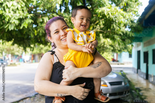 Bild auf Leinwand A Latina mother holding her smiling baby outdoors on a residential street, reflecting love, happiness and family bonding, adult Latina woman and baby 1 to 2 years old