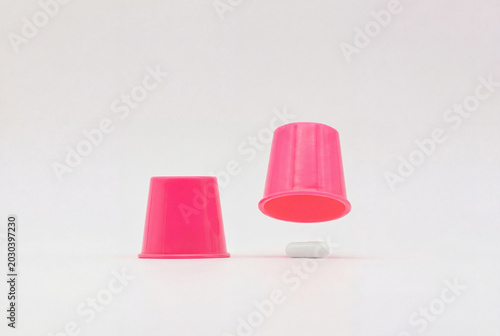 Two pink plastic cups arranged like a shell game with a white capsule revealed underneath, conveying medicine, choice, deception, placebo and risk, in a minimalist bright studio composition.