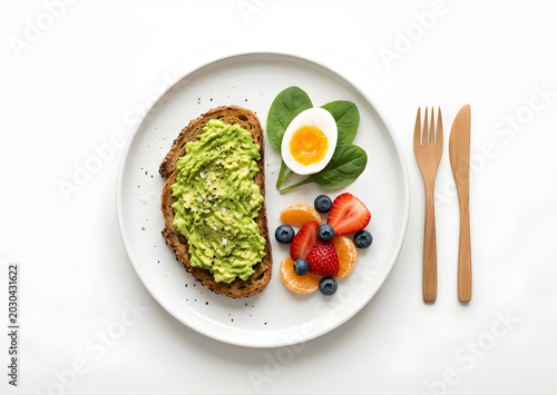 Avocado toast on white plate. Half-hard-boiled egg beside spinach leaves. Fresh berries, sliced strawberries, blueberries, orange. Wooden fork and knife beside plate. Minimalist white background