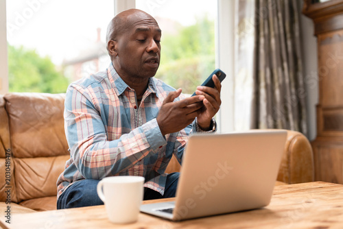 Man using phone while sitting on couch next to laptop with coffee cup on table during daytime at home