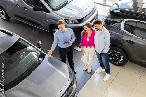 Overhead view of salesman presenting new vehicle options to young couple at modern car dealership showroom during purchase consultation