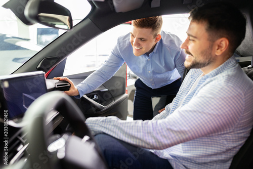 Car salesman showing vehicle interior touchscreen dashboard to smiling male customer seated inside car at dealership. Friendly consultation on features during showroom visit