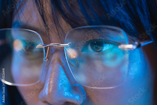 Extreme macro close up of a young Asian woman eye behind a modern rimless eyeglass lens with cool blue light highlighting the iris and her smooth skin texture with a soft cool blue