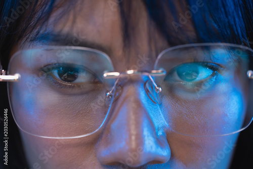 Detailed macro close up of a young Asian woman wearing modern rimless eyeglasses with the lens and frame in sharp focus and cool blue light illuminating her face