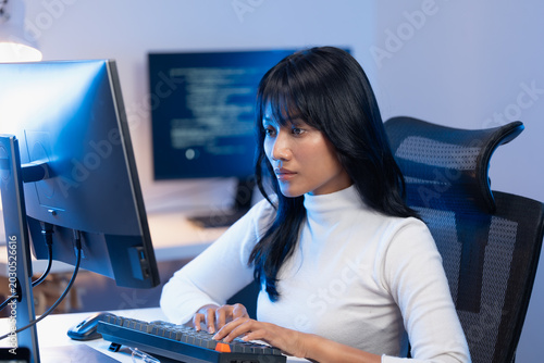 Young Asian woman software developer in elegant white turtleneck typing on her mechanical keyboard at a modern workstation with dual monitors displaying code at night