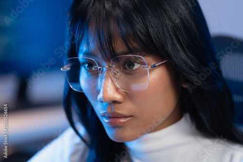 Close up portrait of a confident young Asian female software developer wearing modern rimless eyeglasses and white turtleneck looking sideways in a dark blue lit office