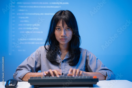 Confident young Asian woman software developer in a blue shirt focused on typing program code on her mechanical keyboard with bright lines of code projected on the wall.