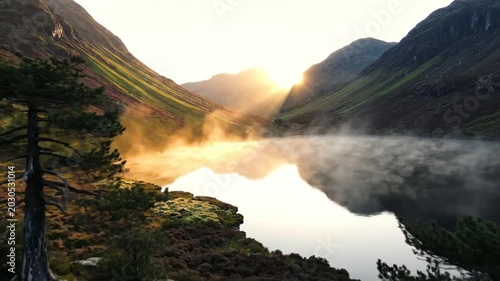 Stunning sunrise over misty lake nestled in a green valley between majestic mountains with sunrays illuminating the scene
