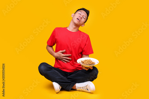 Tableau sur toile Young Asian Man Rolling His Eyes and Holding His Stomach in Pain while Holding P