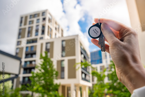 Hand holding house keys with Rental Property keychain near modern apartment building. Real estate income and housing investment concept