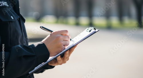 A focused security officer, dressed in a black uniform, takes notes on a clipboard while surveying the area.