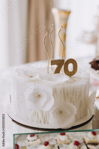 An elegant white birthday cake decorated with delicate edible flowers and a golden number 70 topper, served at a luxury anniversary celebration.