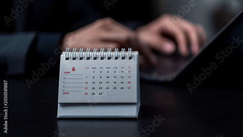Business project planning and schedule management deadline concept. Desk calendar showing the month of June 2026 with a blurred businessman typing on a laptop in the background.
