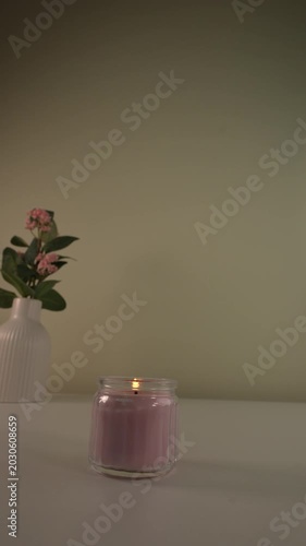 Burning candle and pink flowers bouquet in ceramic vase on white table.