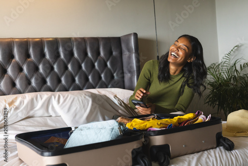Vászonkép Woman sitting on bed holding smartphone and packing carry-on suitcase with straw
