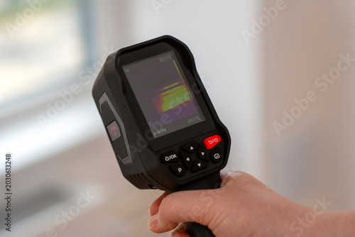 Hand holds a black thermal imager. Its screen shows a color gradient heat map. Red and blue tones indicate temperature differences. The device has 'ON/OFF' buttons and a red button