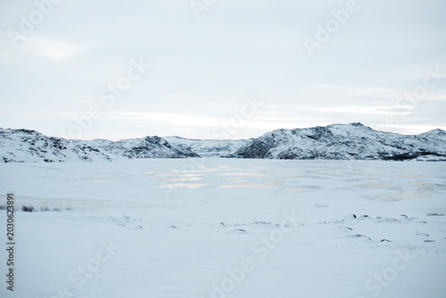 Horizontal shot of frozen lake surrounded by snow covered mountains in Teriberka, winter arctic tundra landscape, cold northern nature wilderness background, polar day white scenic view