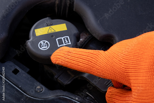 A mechanic uses a gloved hand to point at the antifreeze tank cap on a vehicle while performing maintenance in a garage
