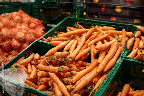 green plastic crates full of loose fresh orange carrots at supermarket, red onions in mesh net bag visible at left, apple display and fruit section behind