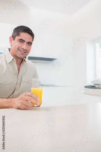 Man sitting at pale kitchen counter holding glass of orange juice, wearing beige shirt, copy space