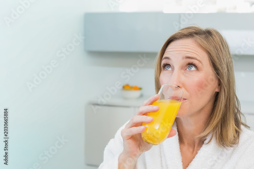 Mature woman wearing white bathrobe sipping from clear glass of orange juice in bright kitchen