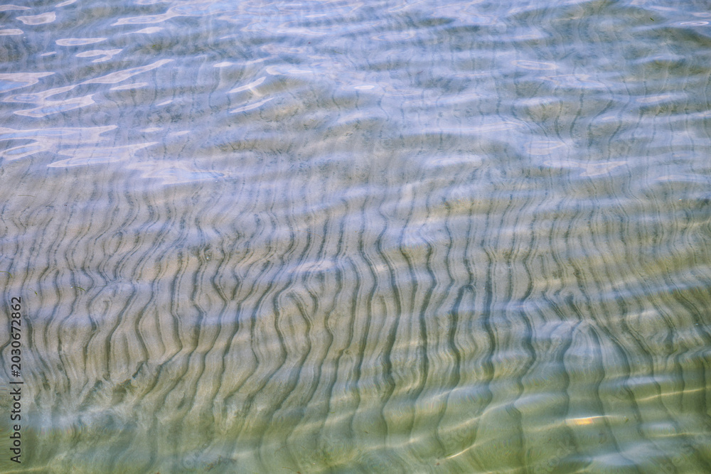 Fototapeta premium gentle ripples on the surface of crystal clear shallow water reveal a textured sandy seabed below.