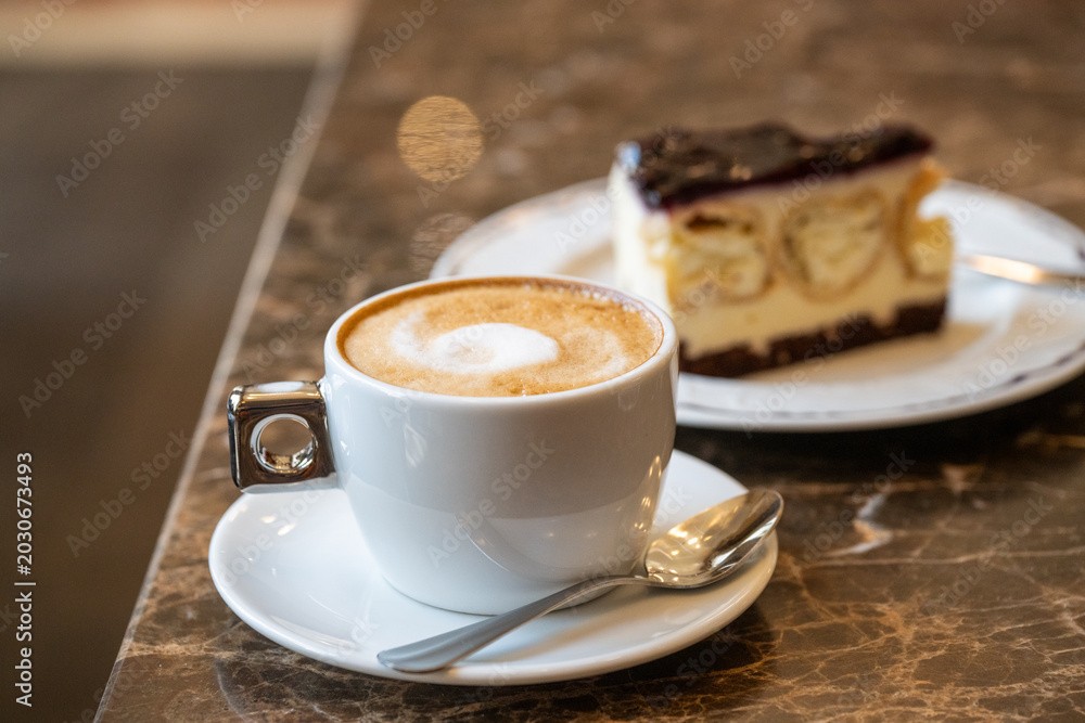 Obraz premium A warm cup of frothy cappuccino sits on a dark marble cafe table alongside a delicious slice of berry-topped cheesecake.
