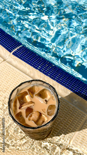 Iced coffee drink with ice cubes in a glass placed beside a swimming pool with clear blue water reflecting sunlight on a warm day
