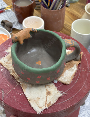 A handmade ceramic cup with a green rim and red stars. It sits on a pottery wheel surrounded by art supplies and unfinished pottery pieces.