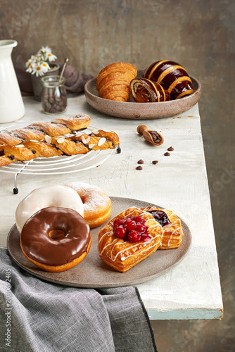 bakery assortment, featuring glazed chocolate and vanilla donuts, cherry and blueberry danish pastries on a ceramic plate, alongside chocolate croissants, a braided puff pastry twist.