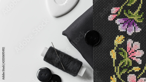 Bild auf Leinwand Black fabric, colorful floral cross-stitch embroidery, a spool of black thread and needle, black buttons, and white tailor's chalk on a clean white surface