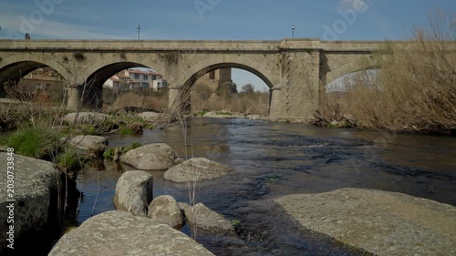 Ancient stone arch bridge spanning a river with rocks and natural vegetation