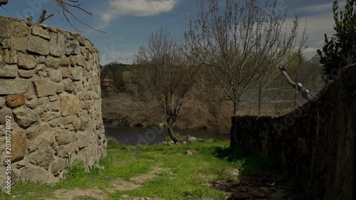 Rustic stone wall guides a dirt path towards flowing river water and trees on...