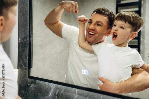 Fotografie Father and son flexing muscles in front of a bathroom mirror, both wearing white