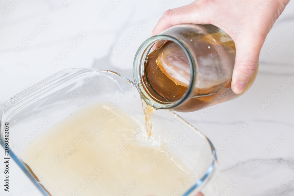 Φωτογραφία Hand pouring amber kombucha tea from glass jar into shallow dish for fermentatio